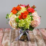 Happy summertime arrangement featuring green and blush hydrangea, orange roses, and variegated orange and blush double petaled roses in a clear base filled with decorative river rocks.