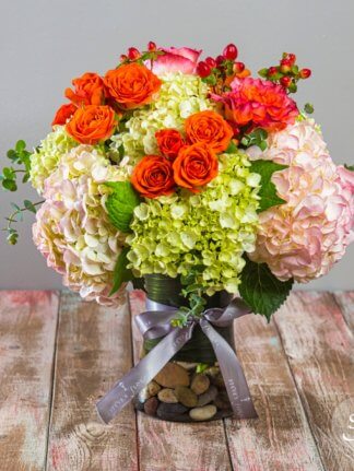 Happy summertime arrangement featuring green and blush hydrangea, orange roses, and variegated orange and blush double petaled roses in a clear base filled with decorative river rocks.